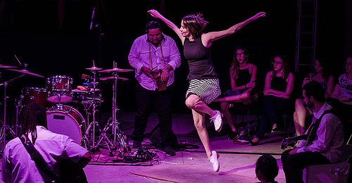 Color photo of Renata, the cast dancer, wearing a horizontal black and white striped skirt, black blouse and white shoes. She jumps leaving the toes of her right foot on the floor, draws in her left leg and opens her arms. All around musicians and audience watch.